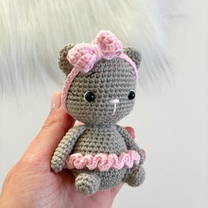 May include: A gray crocheted stuffed animal with a pink bow and a pink tutu. The animal has black eyes and a white nose.