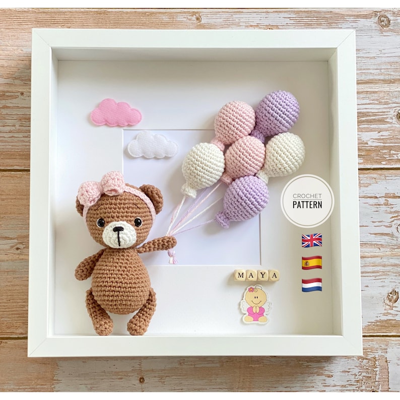 May include: A brown crocheted teddy bear holding pink, white, and purple balloons in a white frame. The frame also includes a white cloud, a pink cloud, and the name "MAYA" spelled out in wooden blocks. The text "CROCHET PATTERN" is also included in the frame.