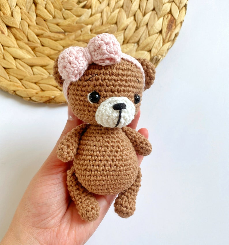 May include: A brown crocheted teddy bear wearing a pink bow. The bear has black eyes and a white nose.