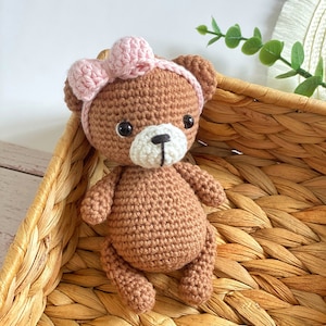 May include: A brown crocheted teddy bear wearing a pink bow headband. The bear is sitting in a woven wicker basket.