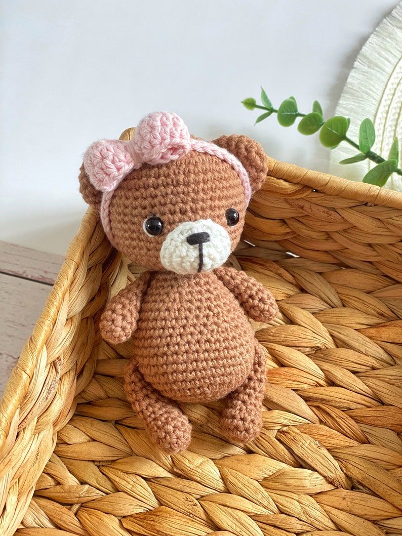 May include: A brown crocheted teddy bear wearing a pink bow headband. The bear is sitting in a woven wicker basket.
