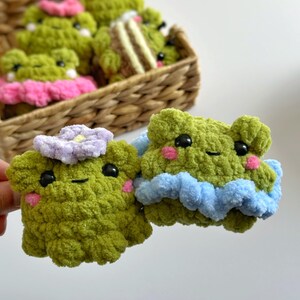 May include: Two handmade, green crochet frog plushies with black eyes and pink cheeks. One frog has a light purple flower, the other has a blue ruffle. A basket in the background holds more frog plushies with pink and yellow accents.