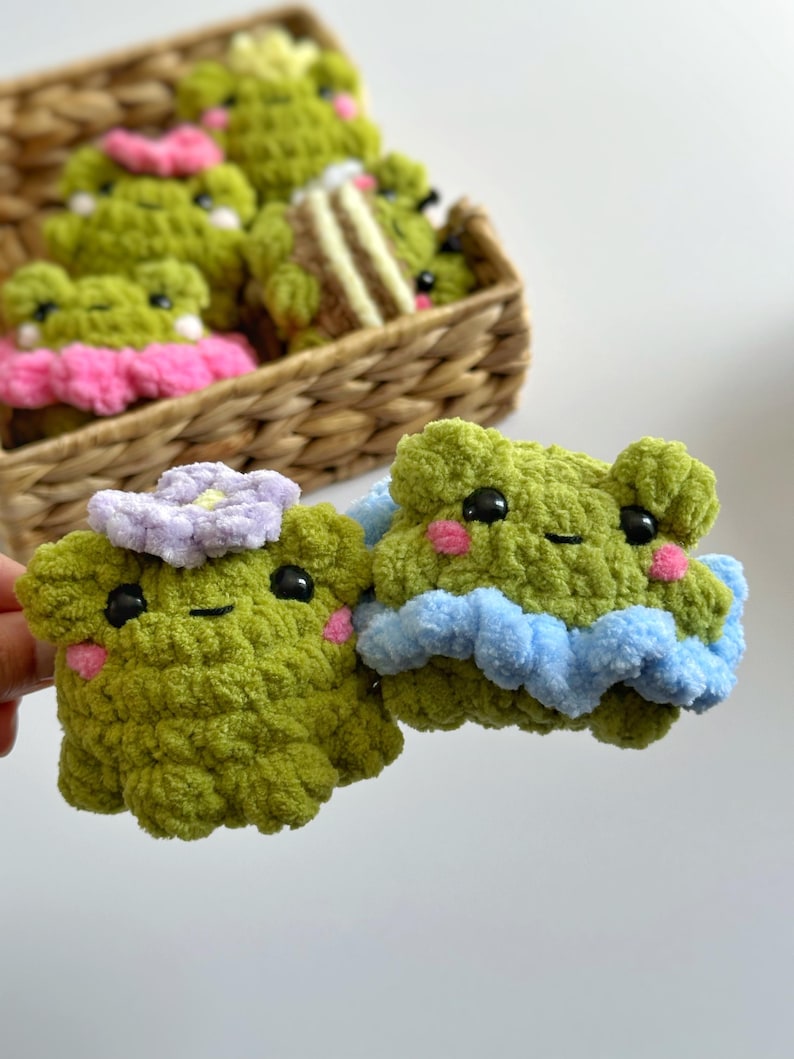 May include: Two handmade, green crochet frog plushies with black eyes and pink cheeks. One frog has a light purple flower, the other has a blue ruffle. A basket in the background holds more frog plushies with pink and yellow accents.