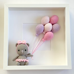 May include: A gray crocheted teddy bear wearing a pink bow and a pink tutu holds a bunch of pink and purple balloons in a white frame.