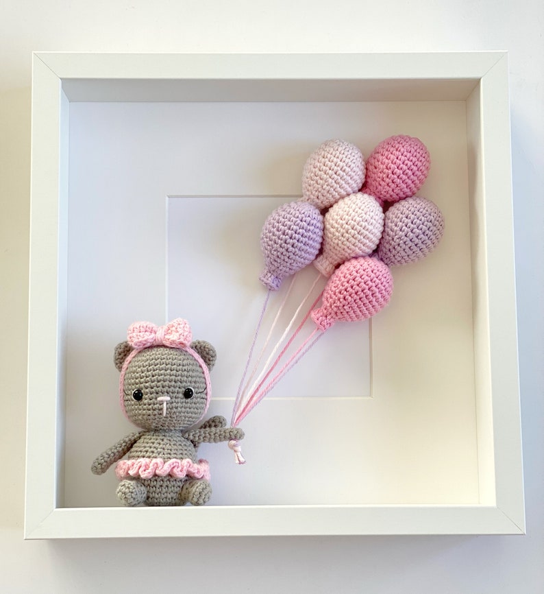 May include: A gray crocheted teddy bear wearing a pink bow and a pink tutu holds a bunch of pink and purple balloons in a white frame.
