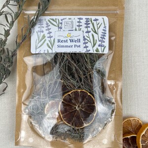 Könnte beinhalten: Ein transparenter Plastikbeutel mit braunem Papierrücken, der eine getrocknete Simmer-Pot-Mischung enthält. Der Beutel hat ein Etikett mit der Aufschrift "Rest Well Simmer Pot" mit einem Lavendel-Blumendesign. Die Mischung enthält getrockneten Rosmarin, Zitronenscheiben und andere Kräuter.