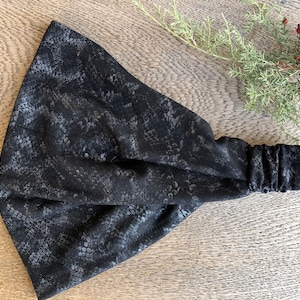 Könnte beinhalten: Ein schwarzes Stirnband mit Schlangenhautmuster. Das Stirnband ist aus weichem, dehnbarem Stoff gefertigt und zum Tragen um den Kopf gedacht.