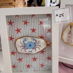 May include: A white framed shadow box containing a painted oyster shell with a blue crab illustration and the word crab written below it. The background features a light blue and white vertical striped pattern with small red starfish icons.