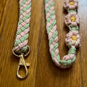 May include: A braided green and pink cord with a gold clasp and pink flower accents. The cord is made of yarn and has a woven design.