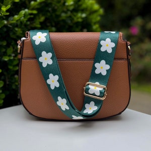 以下が含まれることがあります： 緑と白の花柄の調節可能なストラップが付いた茶色の革製のサッチェルバッグ。ストラップには、濃い緑色の背景に、黄色い中心を持つ白いデイジーが描かれています。