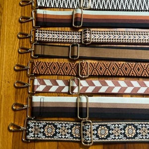 Puede incluir: Ocho correas tejidas de color marrón y blanco con diferentes patrones y cierres de metal dorado. Las correas están dispuestas verticalmente sobre una superficie de madera.