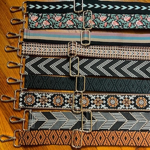 May include: A collection of nine adjustable bag straps featuring various woven patterns including floral motifs, chevron stripes, geometric shapes, and solid bands. Each strap is equipped with gold-toned metal swivel hooks and sliding buckles.