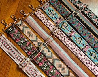 Può includere: Una collezione di nove tracolle regolabili disposte in diagonale. Ogni cinghia presenta motivi distinti, tra cui disegni floreali, forme geometriche e strisce a tinta unita in colori come rosa, blu, verde e marrone, rifinite con ganci girevoli e cursori in metallo dorato.