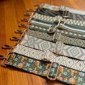 May include: A collection of adjustable bag straps featuring various woven patterns including floral, geometric, and striped designs in shades of green, cream, and blue, each equipped with gold-toned metal swivel hooks and sliding buckles.