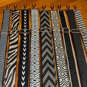 Puede incluir: Una colección de nueve correas ajustables para bolsos con varios patrones en blanco y negro, incluyendo chevron, estampado de cebra, formas geométricas y diseños de grecas, cada una equipada con ganchos giratorios y hebillas deslizantes de metal dorado.