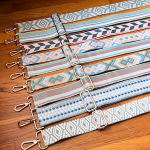 Può includere: Una collezione di tracolle regolabili con vari motivi intrecciati in tonalità di blu, rosa, beige e bianco. Ogni cinghia è dotata di finiture in metallo dorato, tra cui una fibbia scorrevole per regolare la lunghezza e un moschettone.