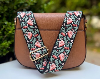 Pode incluir: Uma bolsa de ombro de couro marrom com uma alça estampada de flores pretas e verdes. A alça tem flores rosa e folhas verdes.