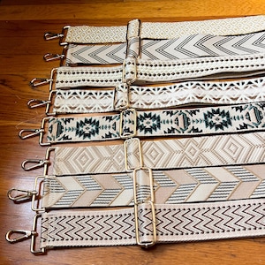 Op de afbeelding: Een collectie van acht verstelbare tasriemen met diverse geweven patronen in neutrale tinten beige, crème, wit en donkergroen. Elke riem is voorzien van goudkleurige metalen hardware, inclusief draaibare karabijnhaken en schuifgespen.