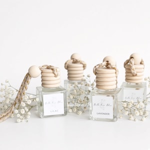 May include: Four square glass car air freshener bottles with wooden caps and braided hanging cords. Each bottle features a white label with the text Bella Terra Bliss and a specific scent name, including Lilac and Lavender, surrounded by baby's breath.