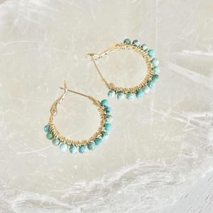 May include: A pair of gold hoop earrings with turquoise beads. The beads are strung along the bottom half of the hoops, which are made of gold-colored wire. The earrings are displayed on a white, marbled surface.
