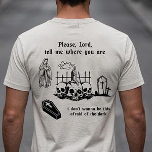 May include: A cream-coloured t-shirt featuring a gothic design. The design includes a coffin, skulls, a candle, a tombstone, and the text "Please, lord, tell me where you are" and "I don't wanna be this afraid of the dark."