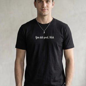 May include: Black crew neck t-shirt with the text "You did good, Slick" in a white gothic font. The person is wearing a silver necklace and a black watch. The background is a plain, light gray wall.