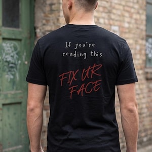 Könnte beinhalten: Schwarzes T-Shirt mit dem Text "If you're reading this FIX UR FACE" in weißer und roter Schrift. Das T-Shirt wird von einer Person getragen, von hinten gesehen, vor einem Hintergrund aus Backsteinmauer.