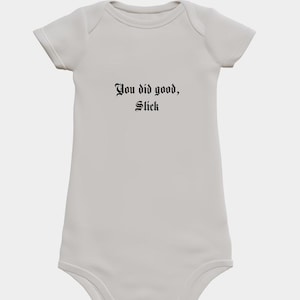 Puede incluir: Un body de bebé gris claro con mangas cortas. El texto "You did good, Slick" está impreso en negro, con una fuente de estilo gótico en el pecho. El body tiene un diseño sencillo y clásico.