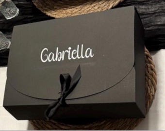 May include: A rectangular black gift box featuring the name Gabriella printed in white script lettering on the front flap. The box is secured with a black ribbon tied into a bow at the center of the curved flap closure, creating a sleek appearance.