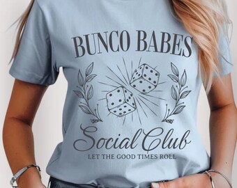 May include: A light blue t-shirt with the text "Bunco Babes Social Club Let the Good Times Roll" and a graphic of two dice with a floral design.