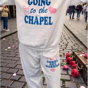 May include: White sweatshirt and sweatpants set with the phrases "GOING to the CHAPEL" and "We're GONNA GET MARRIED" in blue and pink. The outfit is decorated with pink hearts and bows. A wedding theme.