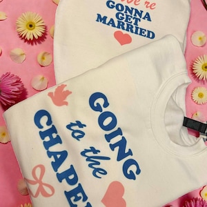 May include: Two white sweatshirts with text and graphics on a pink background with flowers. One sweatshirt says "& We're Gonna Get Married" with a pink heart. The other says "Going to the Chapel" with a pink heart and bow.