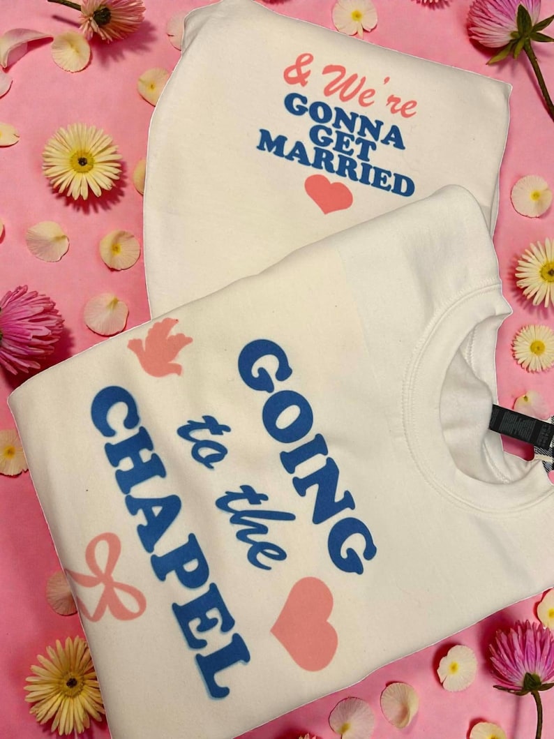 May include: Two white sweatshirts with text and graphics on a pink background with flowers. One sweatshirt says "& We're Gonna Get Married" with a pink heart. The other says "Going to the Chapel" with a pink heart and bow.