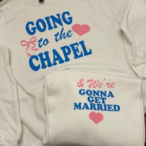 May include: Two white sweatshirts with blue and pink text. One says "GOING to the CHAPEL" with a pink heart and bow. The other says "& We're GONNA GET MARRIED" with a pink heart. Wedding apparel.