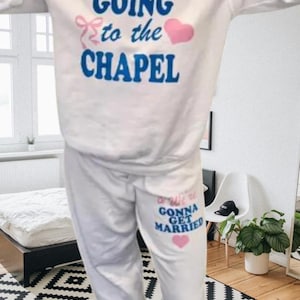 May include: White sweatshirt and sweatpants set. The sweatshirt has the text "GOING to the CHAPEL" in blue with a pink bow and heart. The sweatpants say "We're GONNA GET MARRIED" in blue and pink with a pink heart.