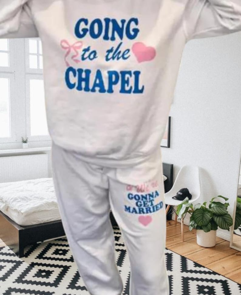 May include: White sweatshirt and sweatpants set. The sweatshirt has the text "GOING to the CHAPEL" in blue with a pink bow and heart. The sweatpants say "We're GONNA GET MARRIED" in blue and pink with a pink heart.