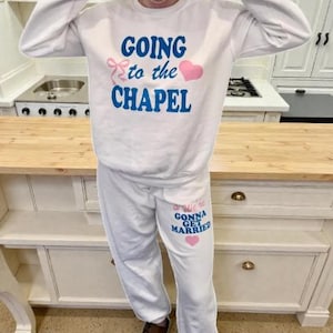 May include: White sweatshirt and sweatpants set with the phrases "GOING to the CHAPEL" and "...We're GONNA GET MARRIED" in blue text, with pink heart and bow accents. The outfit is casual and suitable for a wedding-related event.