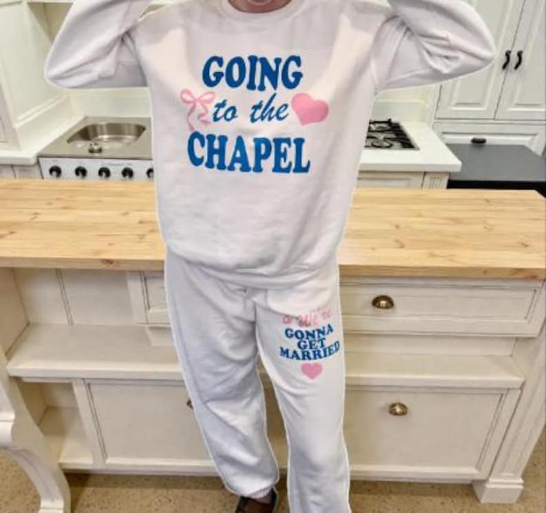 May include: White sweatshirt and sweatpants set with the phrases "GOING to the CHAPEL" and "...We're GONNA GET MARRIED" in blue text, with pink heart and bow accents. The outfit is casual and suitable for a wedding-related event.