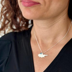 Op de afbeelding: Een zilveren ketting met een hanger in de vorm van Cyprus, versierd met een kleine blauwe edelsteen. De ketting wordt gedragen tegen een zwarte top, waardoor het ontwerp van de hanger wordt benadrukt. De ketting is delicaat.