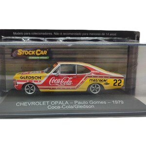 May include: A die-cast Chevrolet Opala model car in a clear display case. The car is yellow and red with Coca-Cola, Gledson, and Metrocar branding, featuring the number 22 on the side. The base is labeled Chevrolet Opala - Paulo Gomes - 1979.