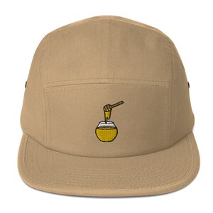 Puede incluir: Una gorra de 5 paneles de color beige con un diseño bordado de un tarro de miel con un cucharón de miel que gotea miel.