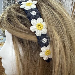 May include: A navy blue headband adorned with crocheted white daisies with yellow centers. The headband is worn on a mannequin head with blonde hair, showcasing the floral accessory.