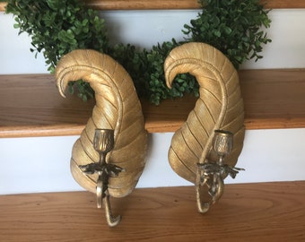 May include: A pair of gold leaf-shaped wall sconces with candle holders. The sconces are made of metal and have a textured finish.