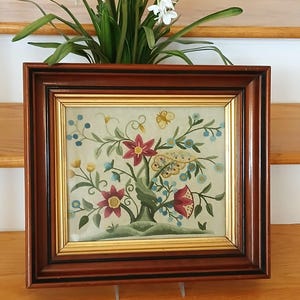 May include: A framed floral embroidery with a dark brown wooden frame and gold inner trim. The embroidery features a tree with red and pink flowers, blue berries, and a butterfly. The frame is sitting on a wooden surface.
