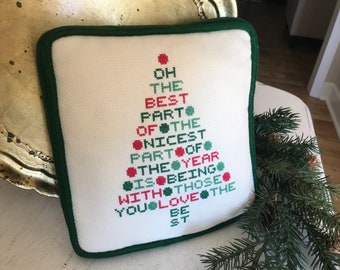 May include: Square, off-white Christmas tree pillow with green trim. The tree design is made of red and green embroidered dots and text that reads "OH THE BEST PART OF THE NICEST PART OF THE YEAR IS BEING WITH THOSE YOU LOVE THE BE ST."