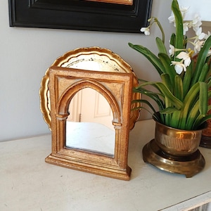 May include: A gold-toned tabletop mirror featuring an arched frame with a rectangular base. The mirror is positioned in front of a larger, round, scalloped-edge gold tray, creating a layered effect with the reflective surfaces.