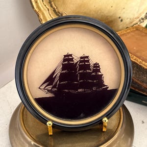May include: A framed silhouette of a tall ship with multiple masts and sails. The black silhouette is set against a tan background within a round, black frame. The frame is displayed on a gold-colored stand.