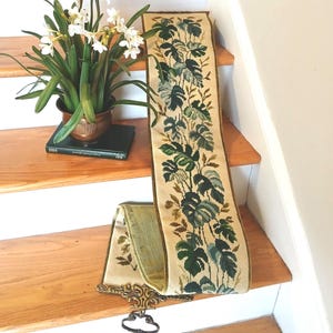 May include: A decorative tapestry runner with a botanical leaf pattern in shades of green and beige. The runner is displayed on wooden stairs, alongside a potted plant with white flowers and a book. A decorative metal piece is attached to the end.