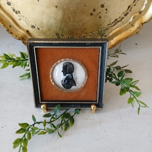 May include: A framed silhouette portrait of a person in profile, set against a rust-colored background. The black and white portrait is in an oval frame, surrounded by a gold-colored border, and encased in a black frame. The frame is supported by small gold-colored stands.