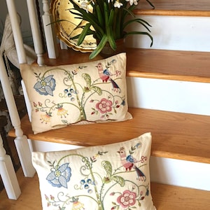May include: Two rectangular cream-colored throw pillows featuring intricate multi-colored embroidery. The design depicts a central tree with stylized flowers, vines, and a bird with blue, red, and pink plumage. The fabric has a smooth, matte finish.